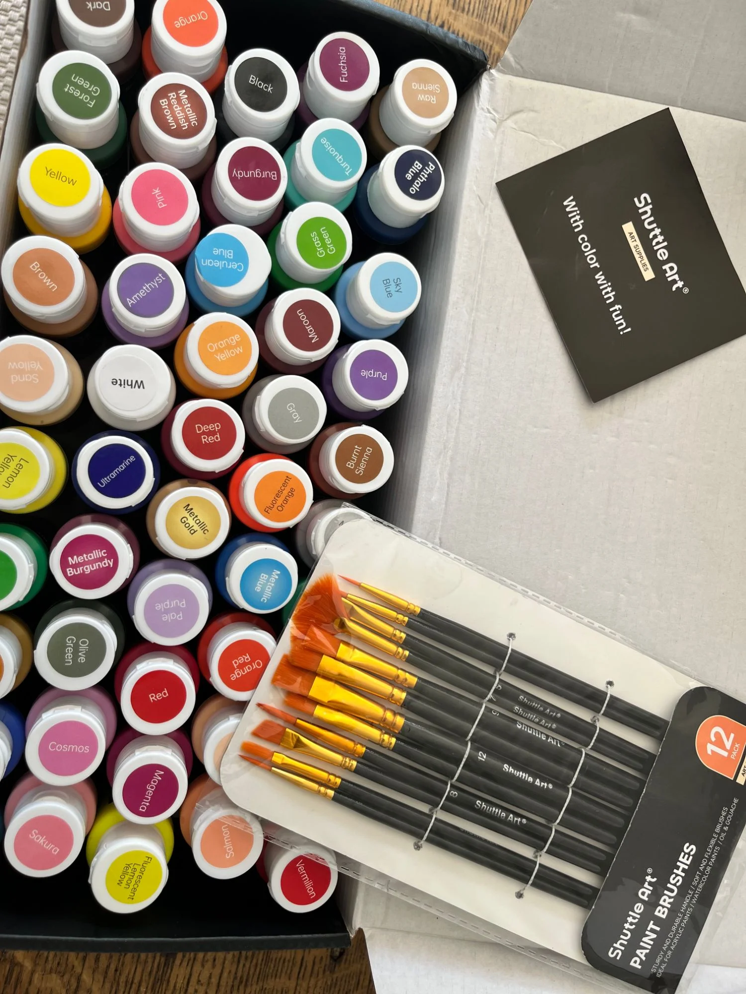The open box seen from above: rows of paint jars with their colour names on the lids, from Forest Green to Vermilion, next to a pack of twelve brushes.
