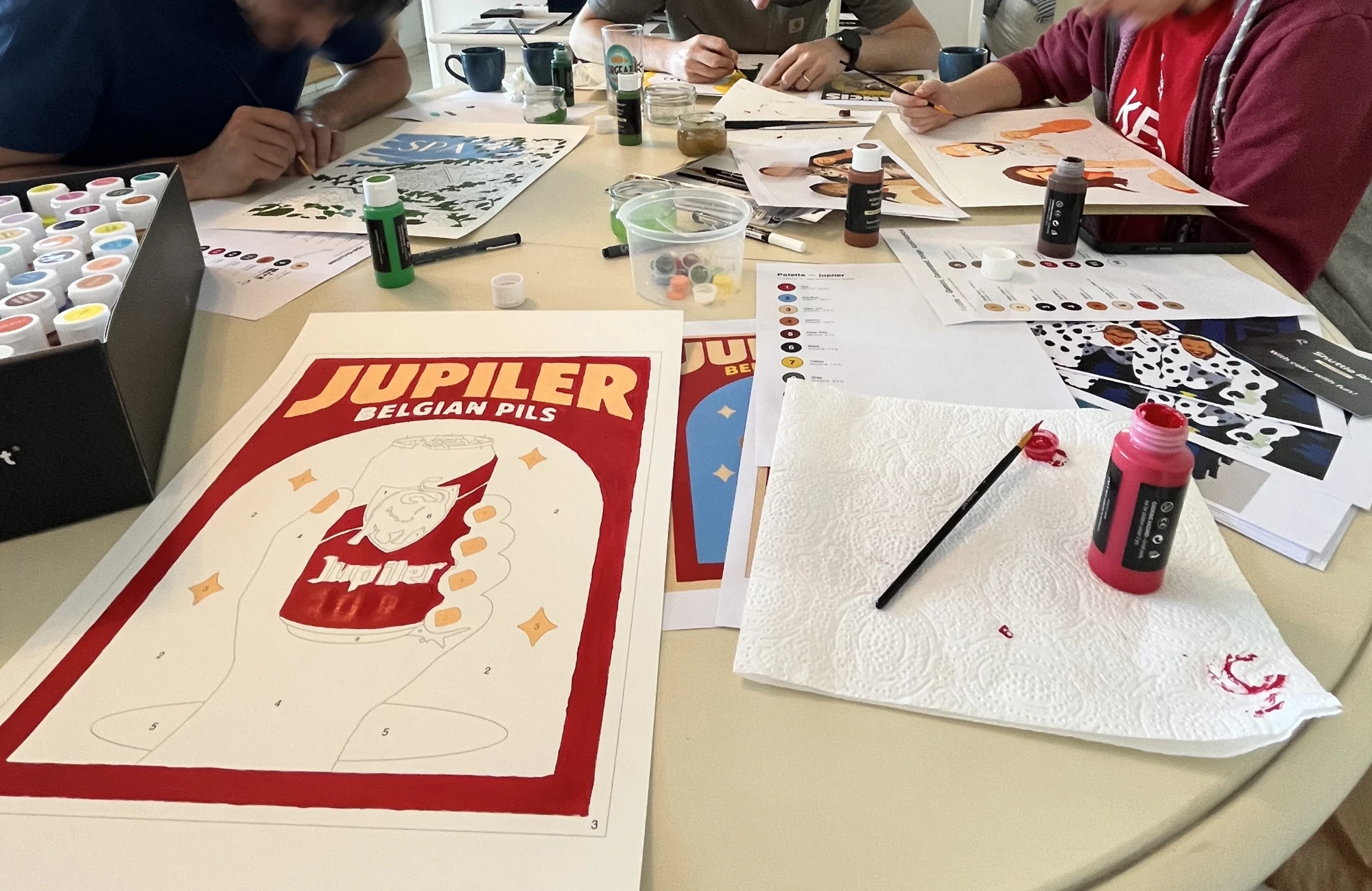Several people painting around a table. In the foreground, the Jupiler poster with its red border and title already painted and the numbered zones of the can still white, next to a jar of red paint, a brush on kitchen paper and the palette sheets.