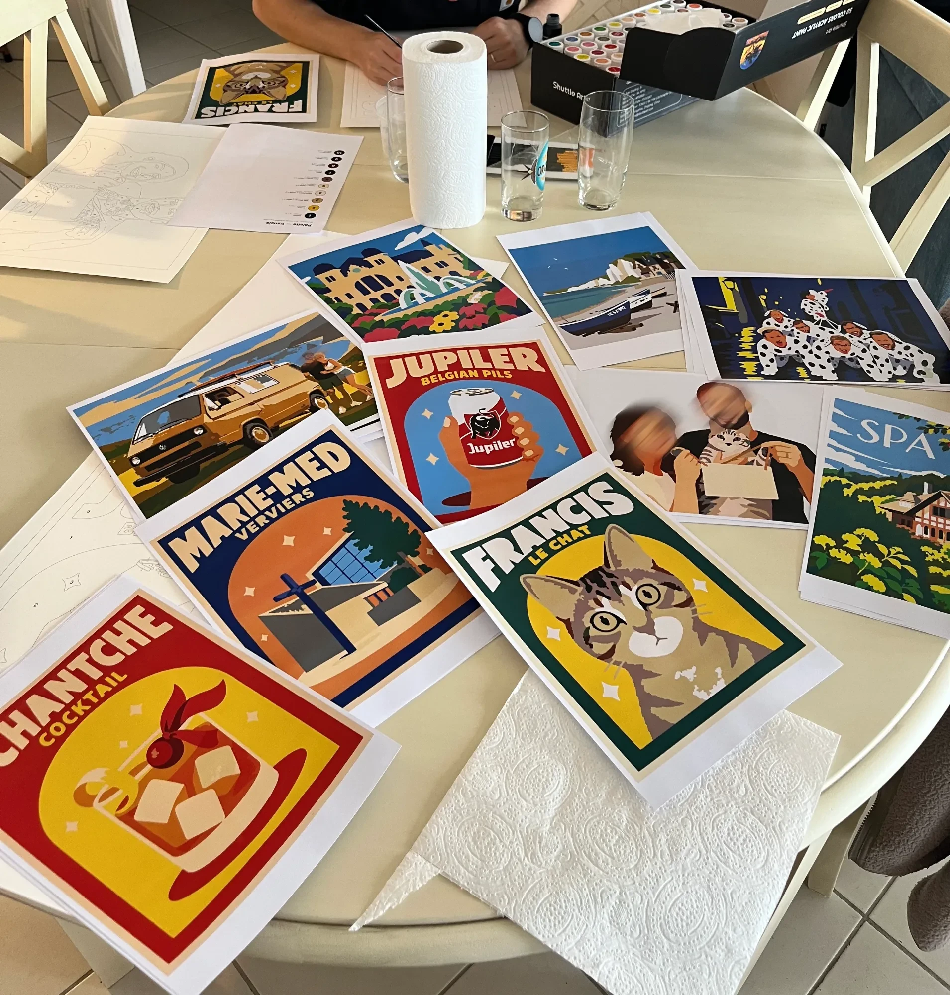 A round table covered with colour prints of the posters, with two line templates and a palette sheet at the back, a roll of kitchen paper, and the open box of paint.