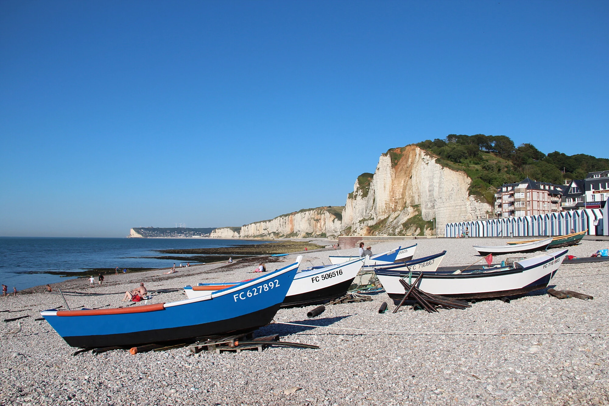 A photograph of the beach at Yport: fishing boats with registration numbers pulled up on the pebbles, bathers, white chalk cliffs and a row of striped beach huts under a clear blue sky.